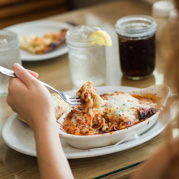 A person's hand uses a fork to lift a cheesy bite of lasagna from a white dish.