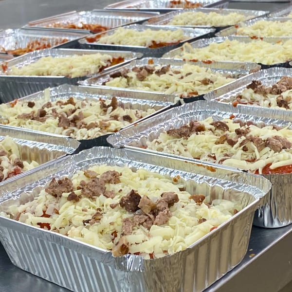 Rows of aluminum trays filled with pasta, sauce, shredded cheese, and crumbled sausage.