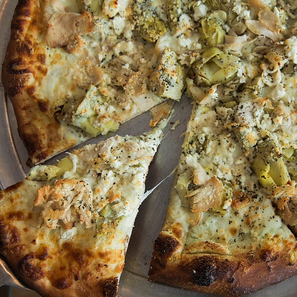 A white pizza from Sally O'Neal's with chicken, artichokes, and feta cheese, with one slice pulled away.