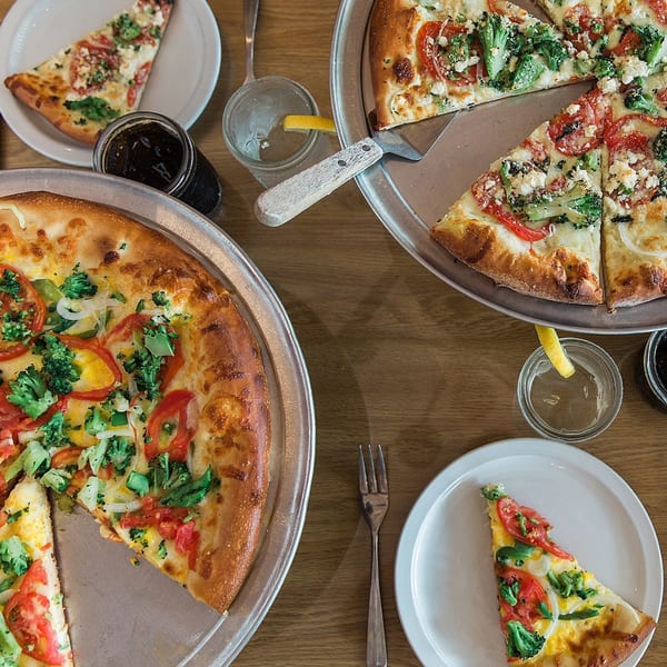 Two large pizzas with broccoli, tomatoes, and cheese, plus individual slices and drinks on a wooden table.