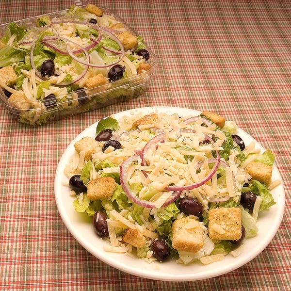 A fresh salad with lettuce, red onion, olives, croutons, and cheese, on a plate and in a to-go container.