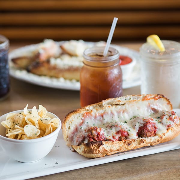 Meatball sub with melted cheese and sauce, a side of chips, and two drinks on a wooden table.