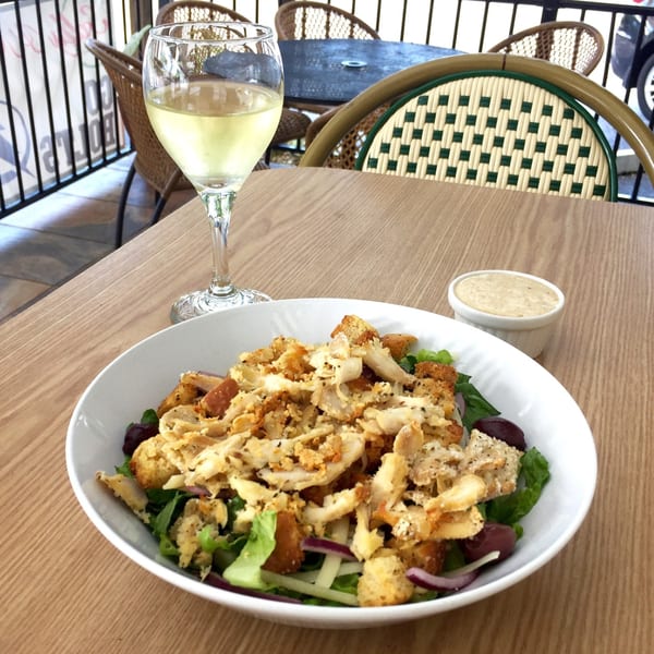 Chicken salad with croutons and olives, a side of dressing, and a glass of white wine on a patio table.