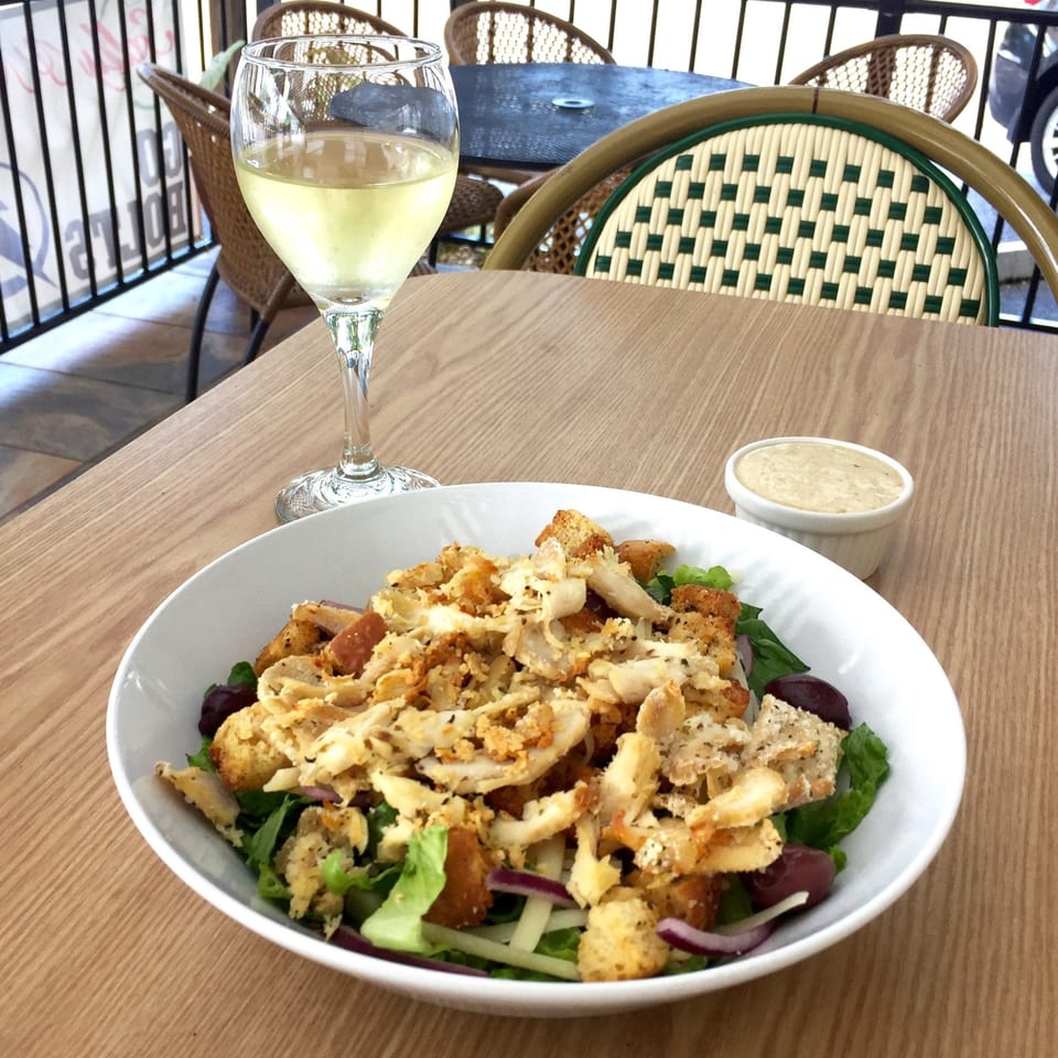 Chicken salad with croutons and olives, a side of dressing, and a glass of white wine on a patio table.
