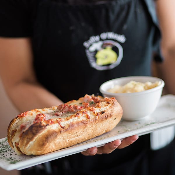 A server in a Sally O'Neal's apron holds a tray with a cheesy baked sub and a side of macaroni.